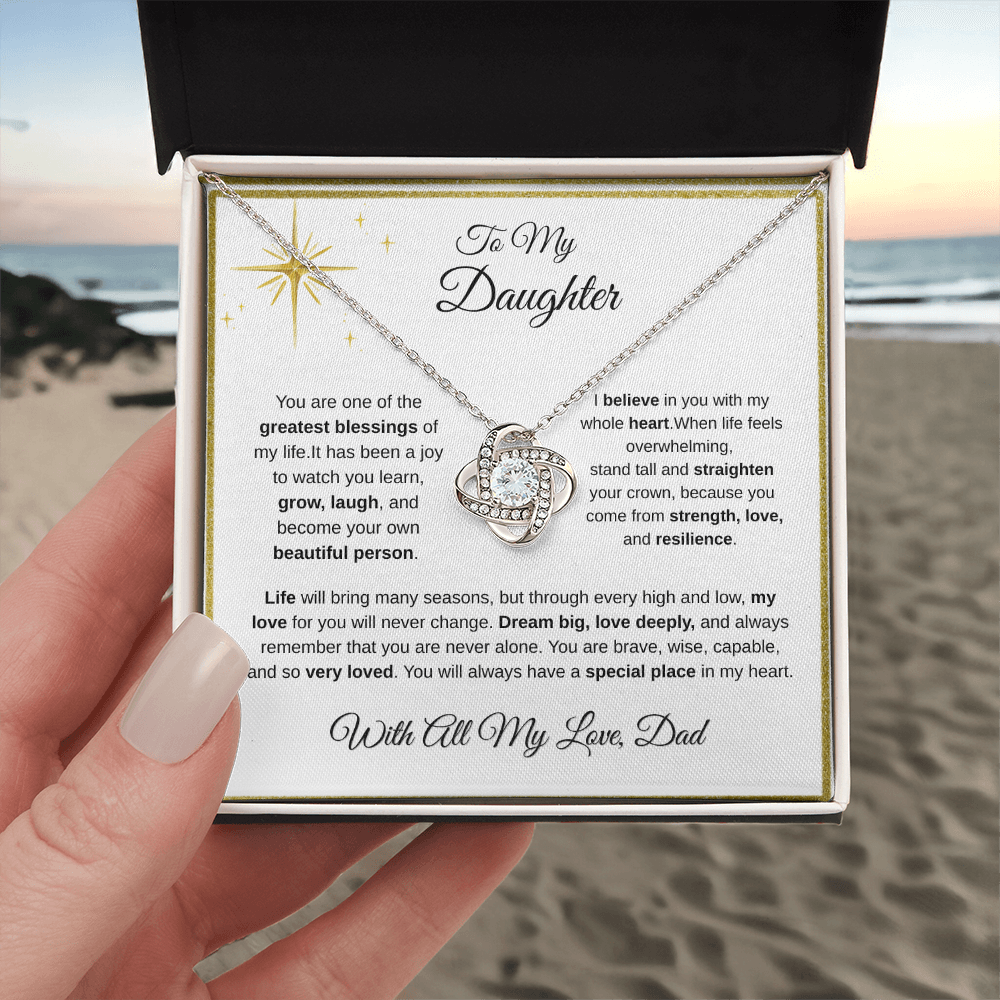 Hand holding 14K white gold-finish Love Knot Necklace in standard gift box at the beach