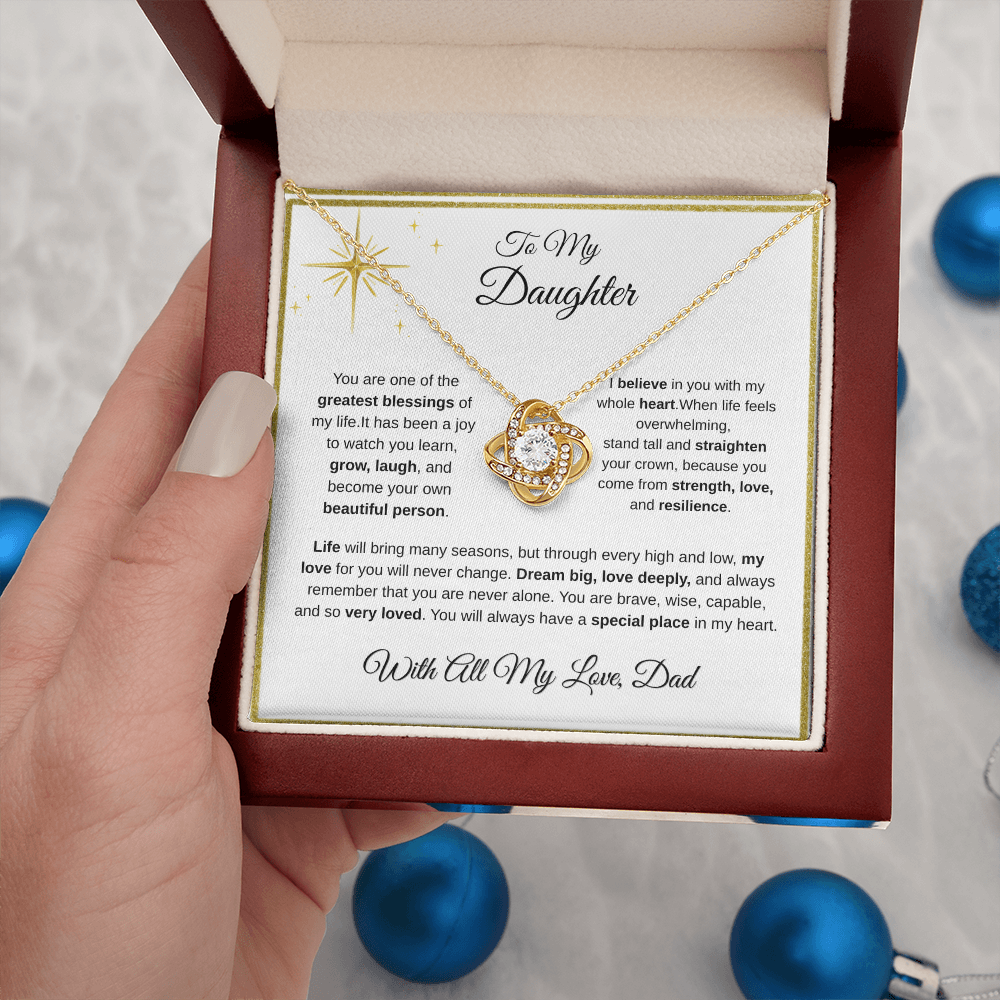 Hand holding 18K yellow gold-finish Love Knot Necklace in mahogany gift box with blue ornaments