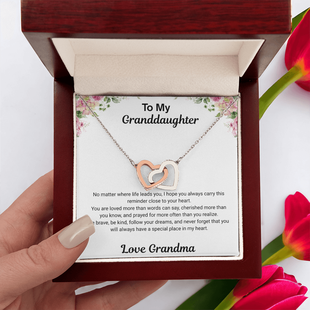 Hand holding 14k white gold-finish Interlocking Hearts Necklace in mahogany-style luxury box with To My Granddaughter message card from Grandma and red flowers